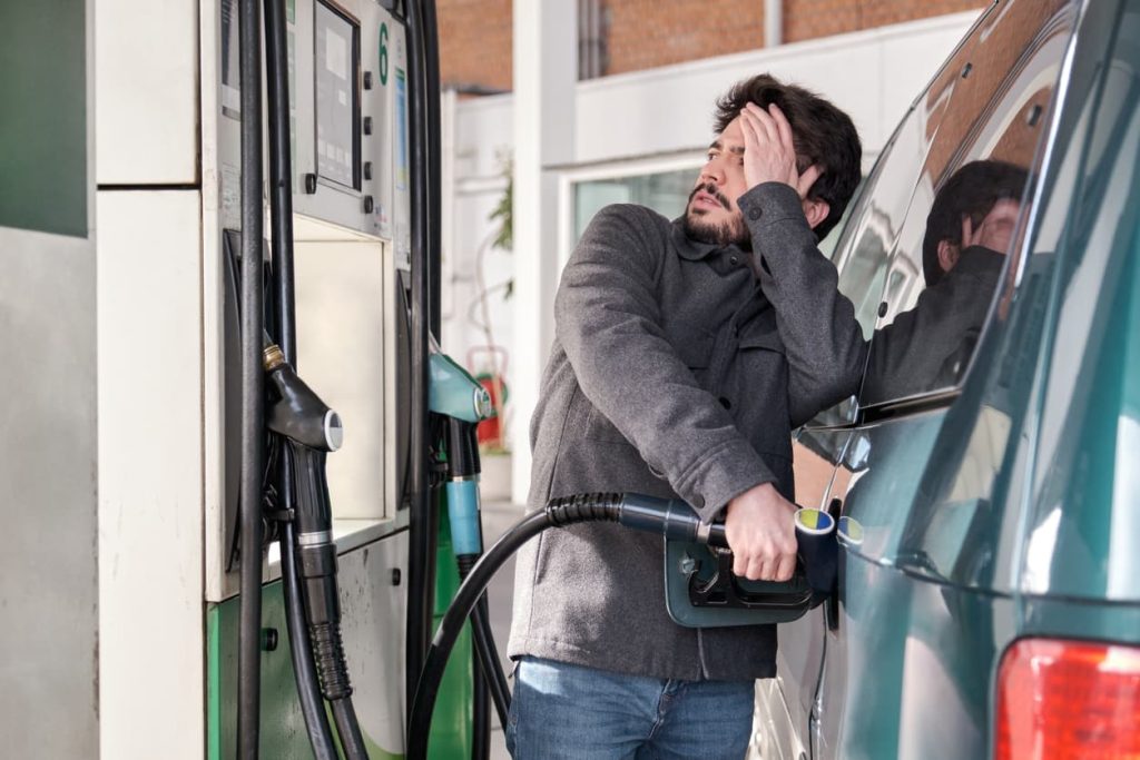 A driver refueling his car while looking distraught at high gas prices at the pump
