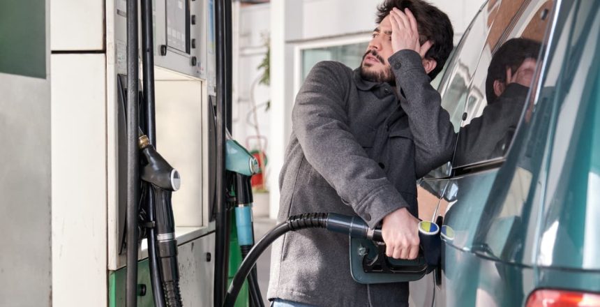 A driver refueling his car while looking distraught at high gas prices at the pump